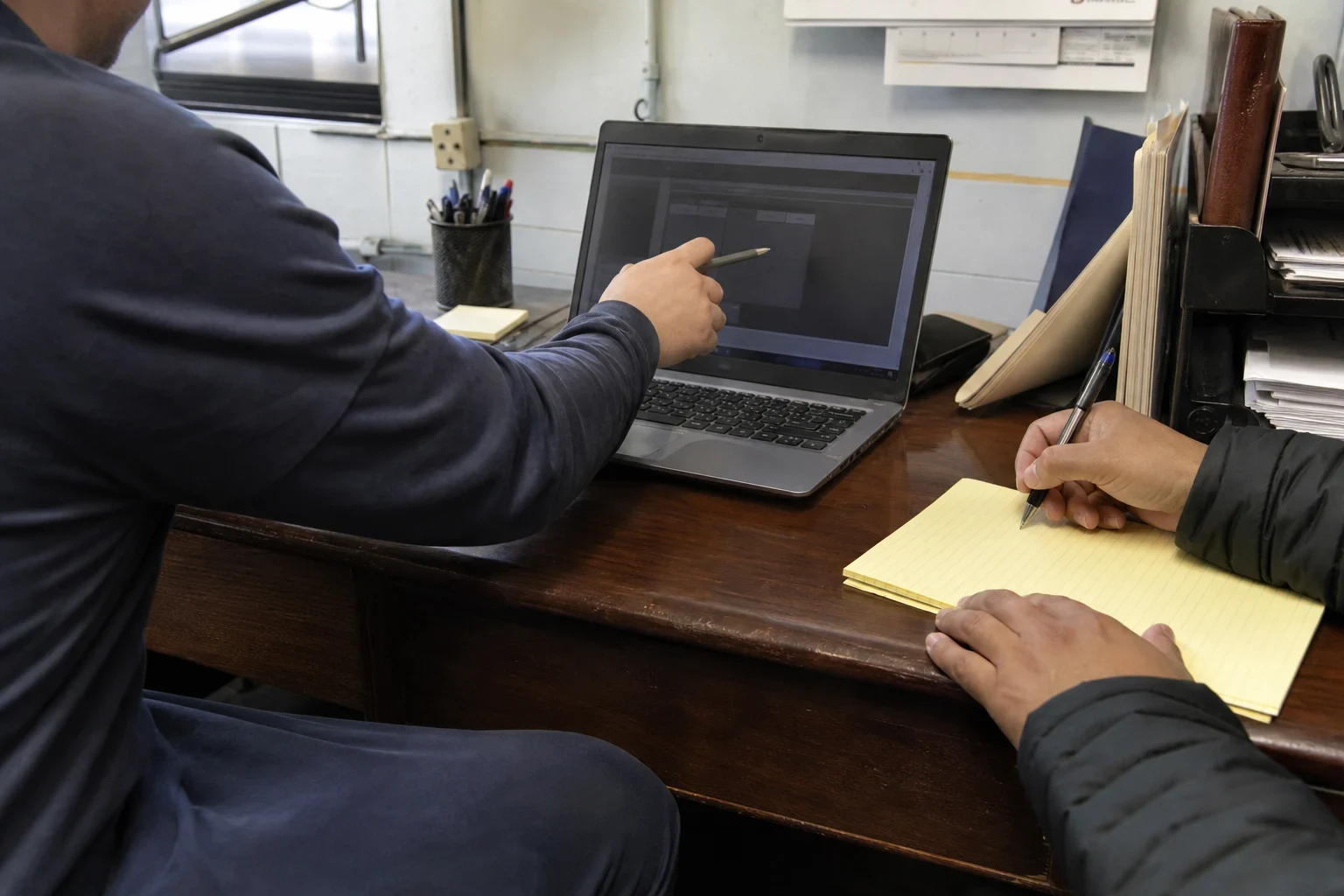 A man points a pen at a laptop screen on a worn wooden desk while another person takes notes on a yellow legal pad in a small office